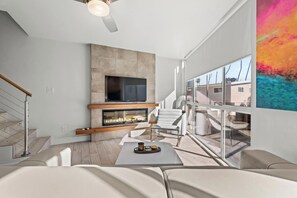 A modern living room with a fireplace, a flat-screen TV, and a large window offering a view of the outside.