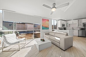 Ein modernes Wohnzimmer mit einem weißen Sofa, einem weißen Couchtisch und einem weißen Sessel. Ein großes Fenster bietet Blick auf einen Pool und einen Balkon.