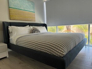A modern bedroom with a large bed, a nightstand, and a window with a view of greenery.