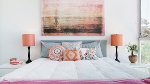 Un dormitorio con cama, almohadas, una mesita de noche, lámpara y ventana.