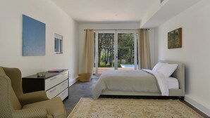 Un dormitori amb un llit, una cadira, un escriptori i un gran finestral amb vista a arbres i una piscina.