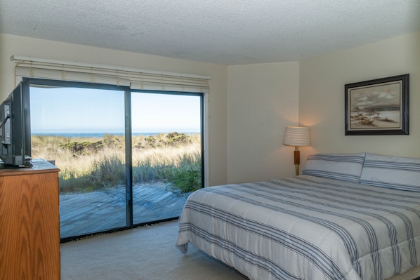 Une chambre avec une grande fenêtre donnant sur la plage et les dunes, un lit avec une literie à rayures, un bureau en bois et une lampe fixée au mur.