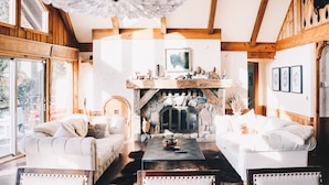 A cozy living room with a fireplace, wooden beams, and large windows.