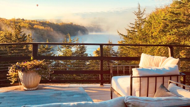 A balcony with a view of a forested area and a body of water. There is a chair and a potted plant on the balcony.