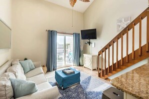 Una sala de estar con sofá, una mesa de centro, televisor, escalera y vistas a través de un ventanal.