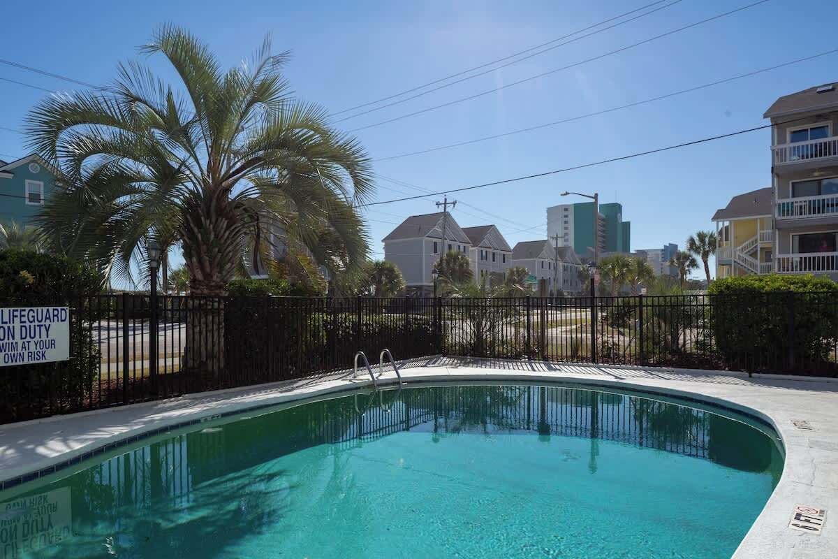 A swimming pool with a lifeguard duty sign, surrounded by a fence and palm trees.