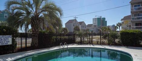 A swimming pool with a lifeguard duty sign, surrounded by a fence and palm trees.