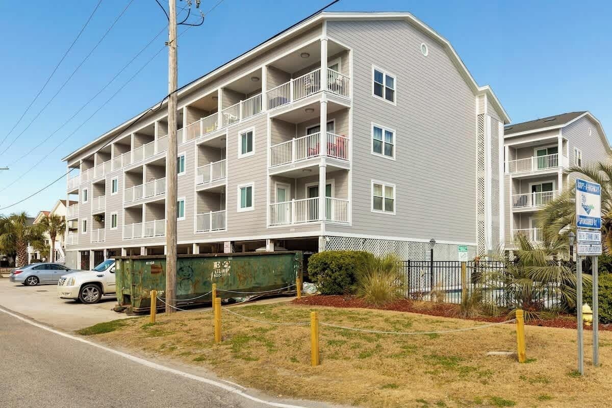 A multi-story residential building with balconies, a gated parking area, and a clear sky.