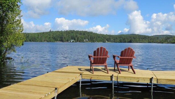 Puinen laituri, jolla on kaksi punaista Adirondack-tuolia, ja ne ovat käännettyinä kohti tyyntä järveä. Laituria ympäröi tiheä metsä ja sen yllä on kirkas taivas.