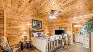 A wooden cabin bedroom with a bed, rocking chair, bedside table, lamp, and a ceiling fan.
