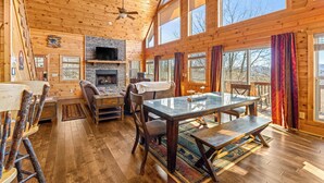 A spacious wooden cabin interior with a large dining table, a fireplace, and multiple windows offering views of the outdoors.