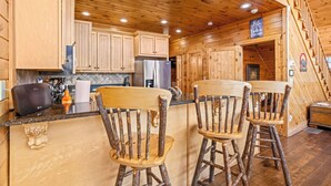 A rustic kitchen with wooden cabinets, a refrigerator, and a bar area with two wooden chairs.