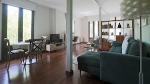 A spacious living room with a green sofa, wooden flooring, a television, and a bookshelf filled with books and decorative items.