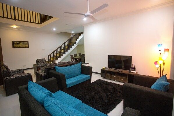 Una sala de estar moderna con una escalera, sofás negros y azules, un televisor de pantalla plana y un ventilador de techo.