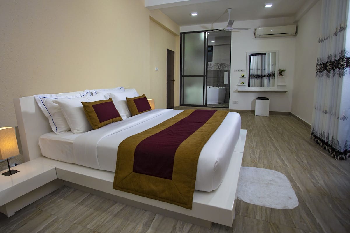 Quarto de hotel moderno com uma cama grande, uma mesa de cabeceira com abajur, uma porta de vidro deslizante para o banheiro e um pequeno armário com produtos de higiene pessoal.
