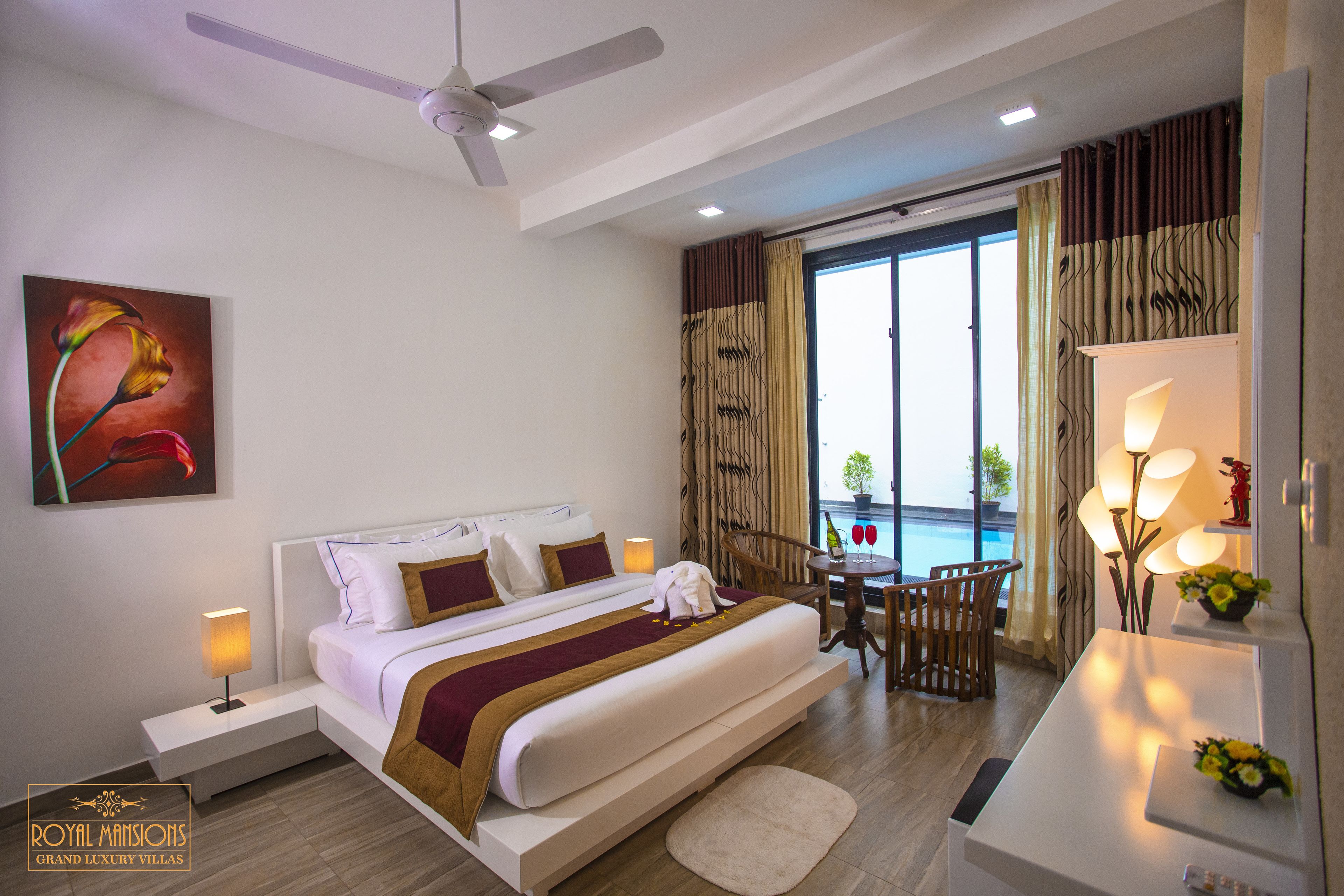 A hotel room with a bed, bedside table, a desk with a chair, a balcony with curtains, and a ceiling fan.