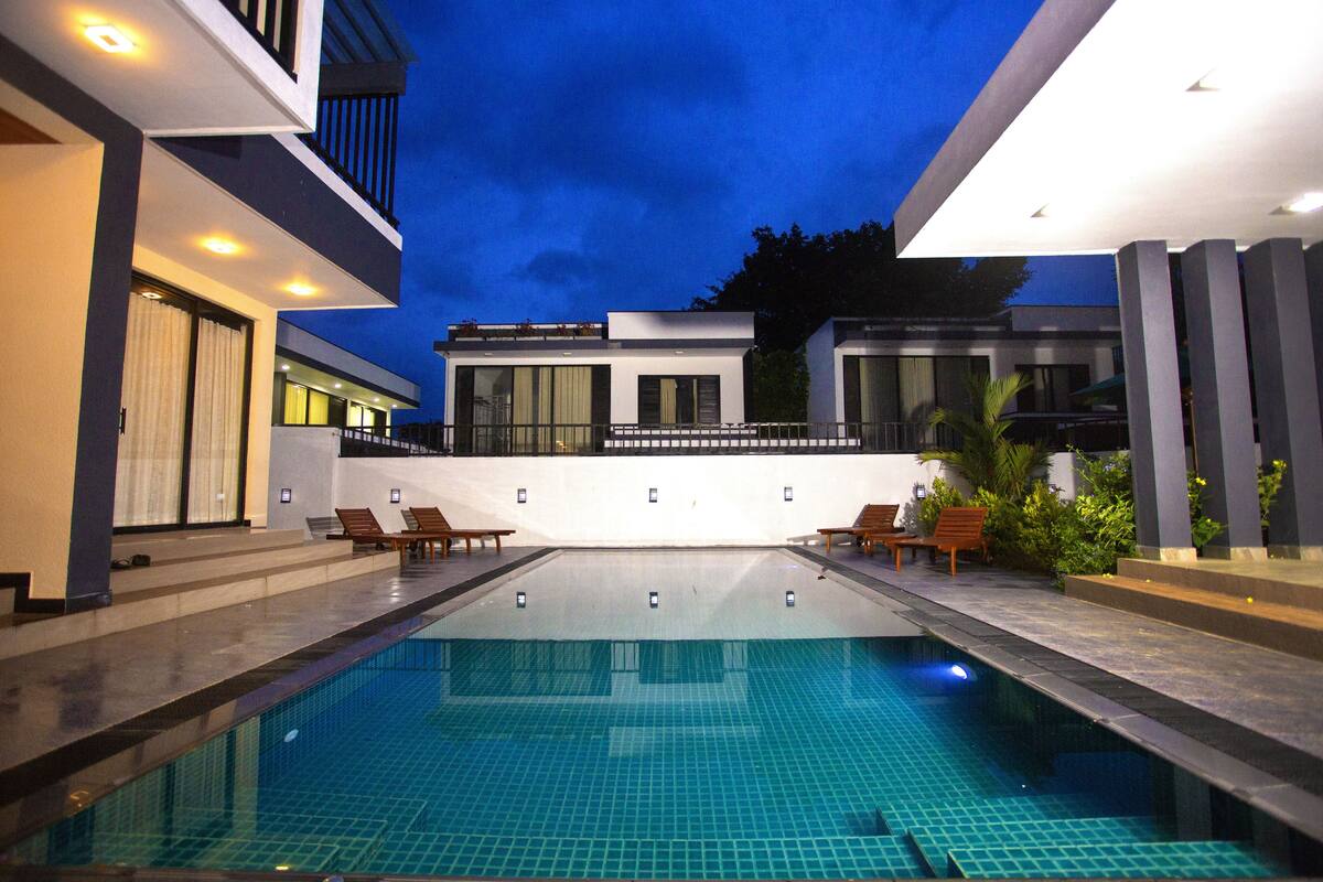 Uma piscina moderna ao ar livre com superfície de água azul cristalina, rodeada por um terraço bem iluminado com espreguiçadeiras e um edifício contemporâneo ao fundo.