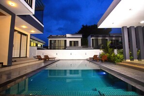 Una piscina exterior moderna con superficie de agua azul cristalina, rodeada por un patio bien iluminado con sillones de descanso y un edificio contemporáneo al fondo.
