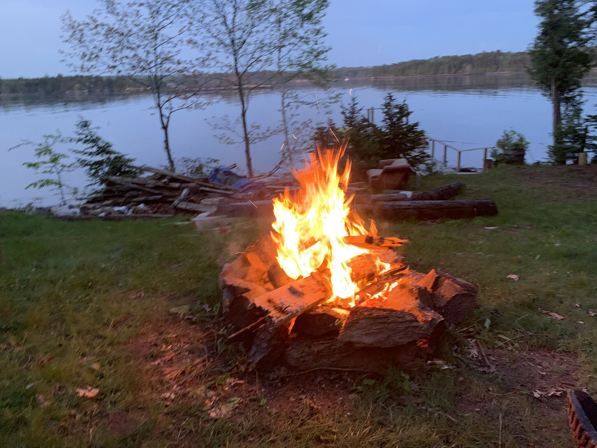 Un feu de camp dans un cadre naturel, avec un lac et des arbres en arrière-plan.