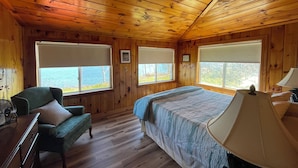 Quarto de cabana com cama, poltrona, escrivaninha e janelas amplas com persianas.