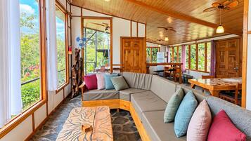 Amplia habitación con un techo de madera, suelo de piedra, un banco de madera, un sofá con cojines y ventanas grandes con vistas a la vegetación.