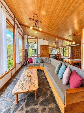 Une pièce spacieuse avec un grand plafond en bois, un sol en pierre, un banc en bois, un canapé avec des coussins et de grandes fenêtres donnant sur un paysage verdoyant.