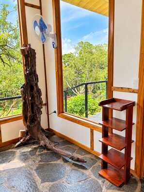 A room with a large window, a wooden sculpture, a fan, and a wooden shelf.
