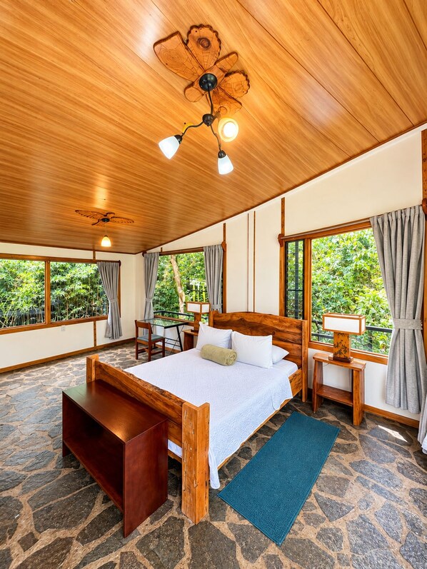 A room with a large bed, a wooden ceiling fan, a small table with a lamp, and a view of greenery through the windows.