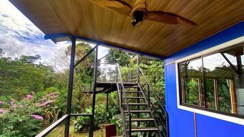 A wooden ceiling fan, a staircase, and a table with chairs on a balcony with a view of a garden.