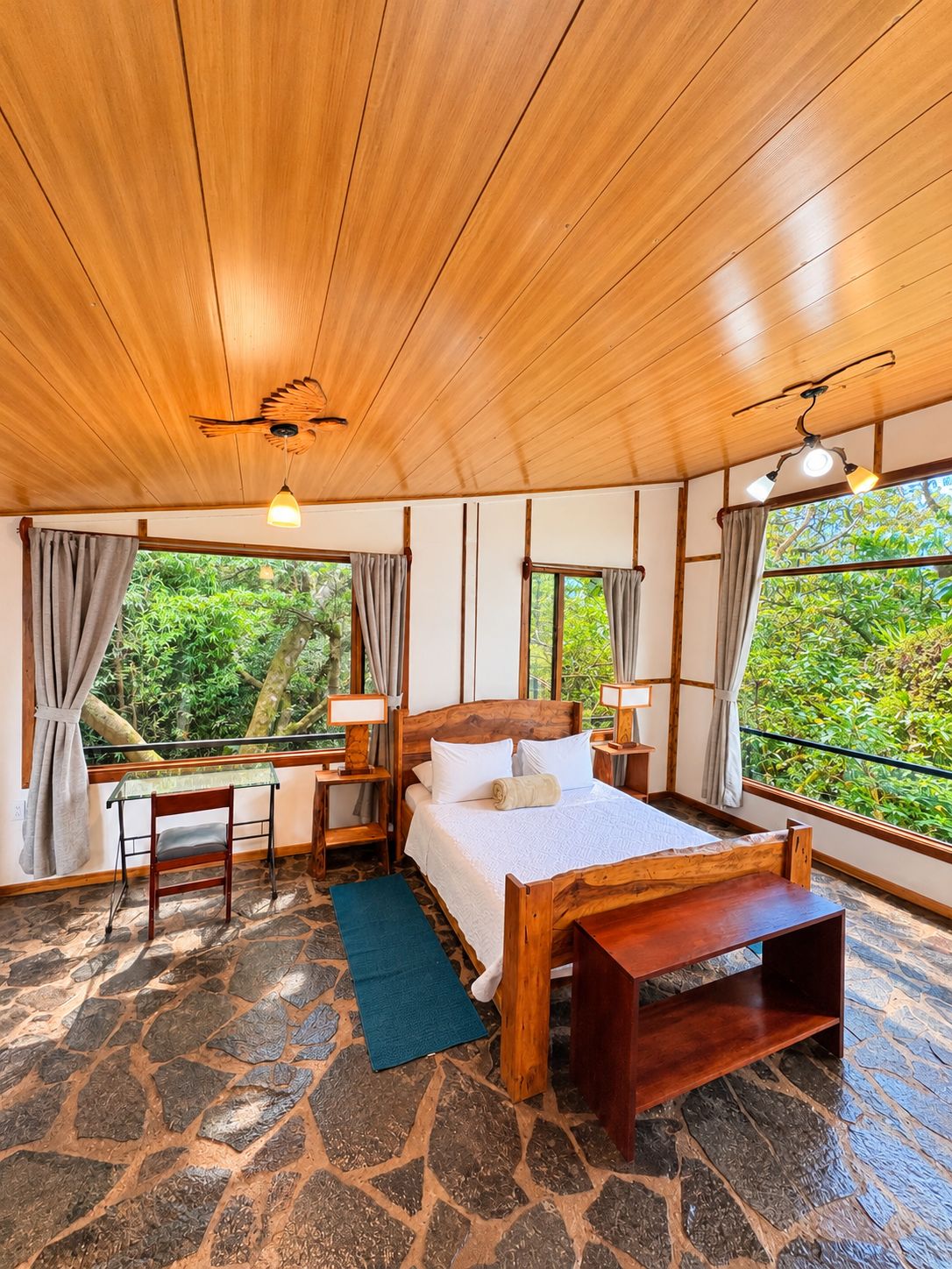 A wooden room with a bed, a desk, and a chair. There are windows with curtains, a ceiling fan, and a view of greenery outside.