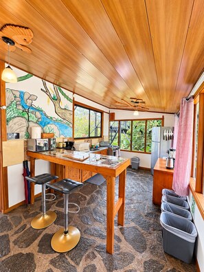 A kitchen with a wooden table, bar stools, a refrigerator, and a window with a view of greenery.