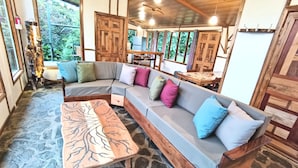 Un intérieur chaleureux et rustique, avec un grand canapé d’angle en bois, un sol carrelé en pierre et une table basse en bois ornée d’un motif d’arbre.