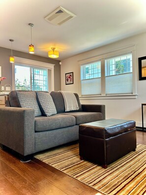 A modern living room with a grey sofa, a brown ottoman, and a striped rug. There are two pendant lights, a ceiling vent, and a window with blinds. - Cozy Private Cottage Near Downtown Portland (Portland)