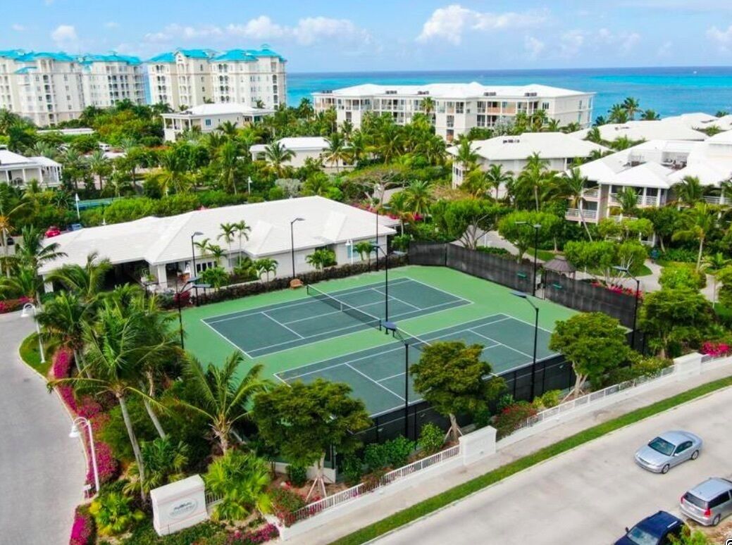 Vista aérea de un resort con cancha de tenis rodeada de abundante vegetación y palmeras, junto a una carretera con coches estacionados.