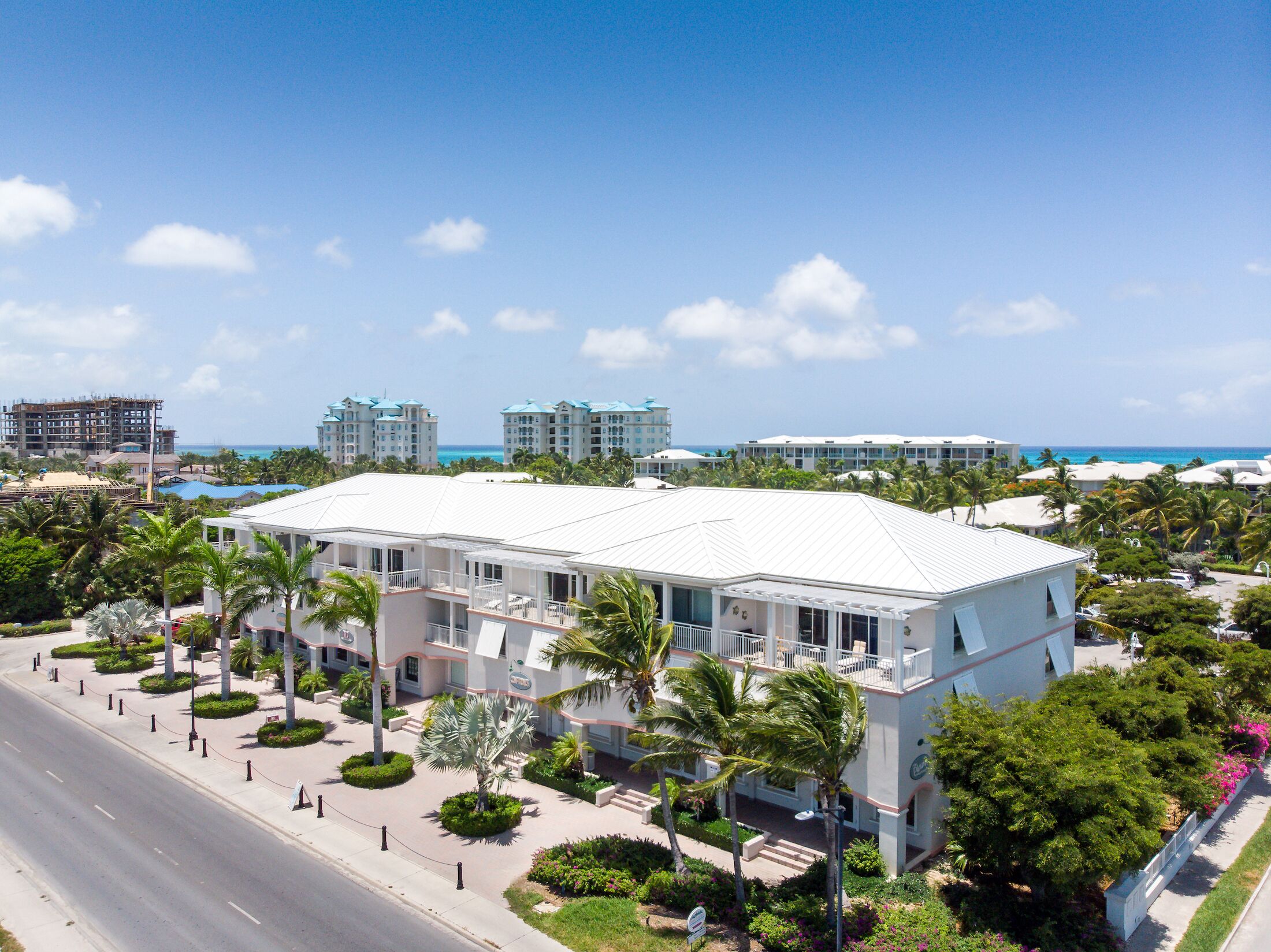 A hotel with a white facade, multiple floors, and a flat roof. It is surrounded by palm trees and has a view of the ocean.