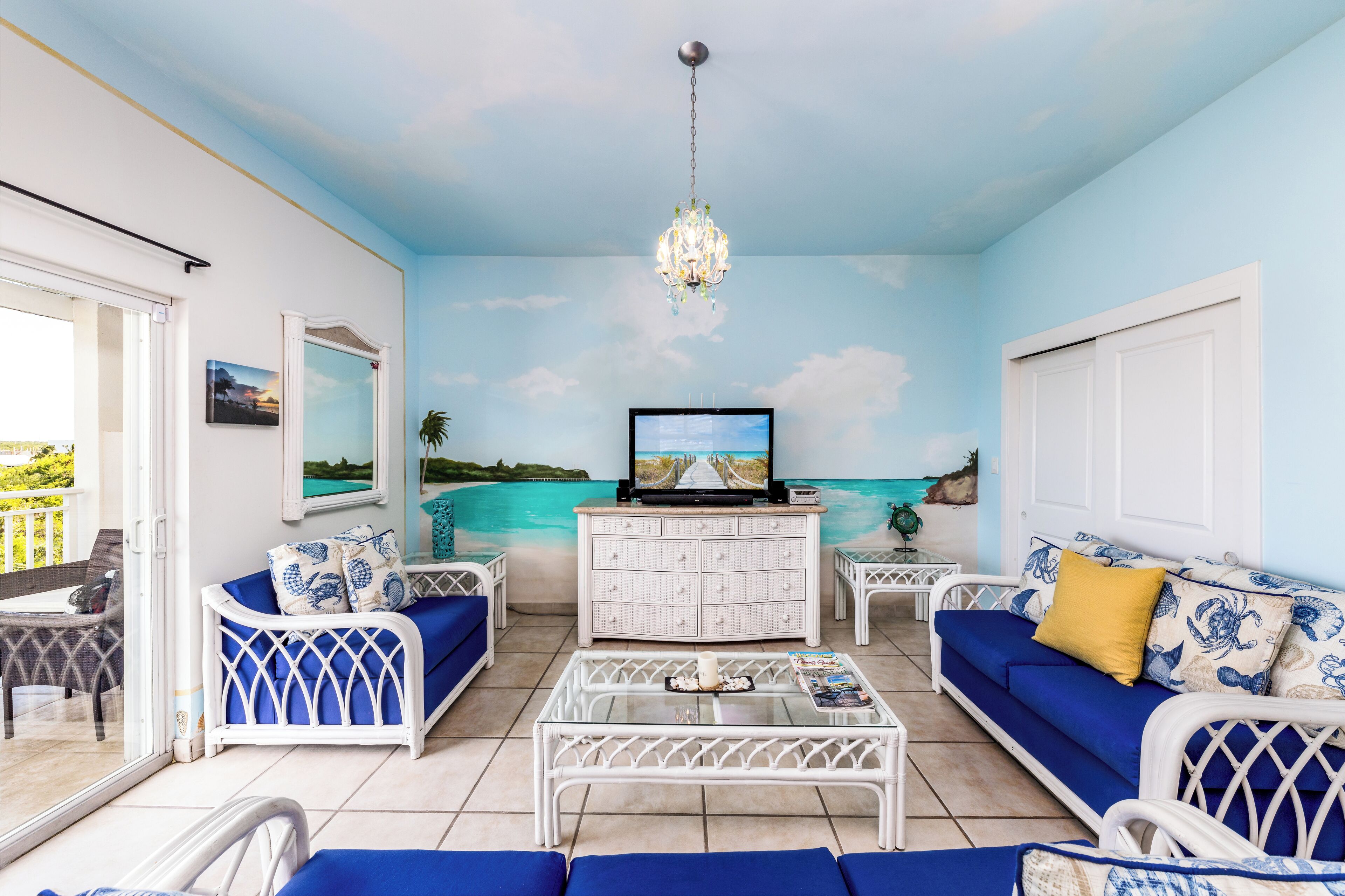 A living room with a TV, white wicker furniture, a glass coffee table, and a mural of a beach scene.