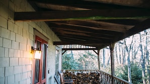 A covered porch with a stack of firewood, a wooden bench, and a wall-mounted light fixture. - Walk to DT Highlands | Designer Cabin | Fireplace | Fenced Yard | Dog Friendly (Highlands)