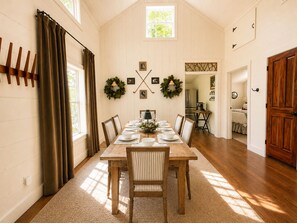 Une salle à manger avec une table en bois dressée pour six personnes, flanquée de deux fenêtres ornées de rideaux, une cuisine en arrière-plan et une porte en bois sur la droite.