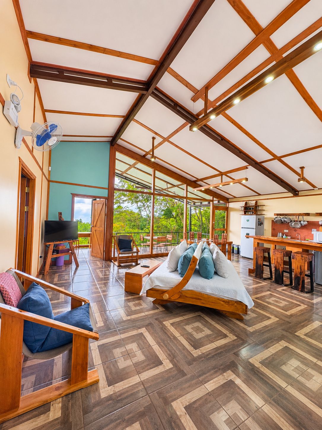 Una habitación amplia con techo de madera, una cama con almohadas, un televisor y un comedor con cocina al fondo.