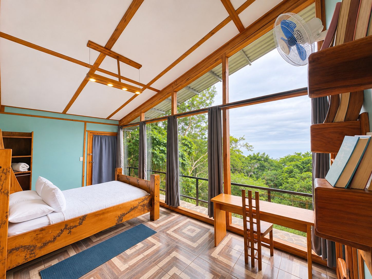 Un dormitorio con cama, escritorio y silla, ventanas amplias con vista a follaje y un ventilador de techo.