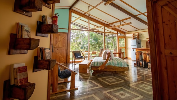 A spacious room with a high ceiling, wooden beams, a bed with a green and white striped bedspread, a wooden chair, and a bookshelf filled with books. - Above the Gulf House: Spectacular Ocean View! (Monteverde)