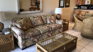 Ein Wohnzimmer mit einer Couch, einem Couchtisch, Korbmöbeln, einer Lampe und einer Küche im Hintergrund.