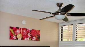 Ein ordentlich bezogenes Bett mit weißen Leinentüchern, ein Deckenventilator, ein Wandbild mit Blumen und ein Fenster mit Jalousien.
