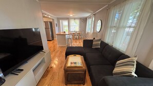 Ein modernes Wohnzimmer mit einem Flachbildfernseher, einem schwarzen Sofa, einem Holztisch und einem Essbereich im Hintergrund.