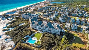 An aerial view of a coastal residential area with a large, multi-story house featuring a swimming pool, surrounded by smaller houses and a beach. - Direct Gulffront! BREATHTAKING 270 degree Ocean Views! Large 2600 sqft 3bdrm! (WaterSound Beach)