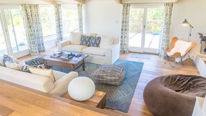 Ein geräumiges Wohnzimmer mit einer Couch, einem Couchtisch und einem Hocker, einem großen Fenster mit Vorhängen und einer Holzbalkendecke.