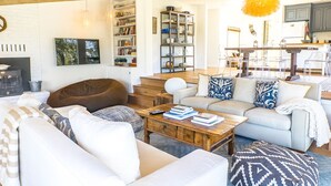 Ein modernes Wohnzimmer mit einer Couch, einem Couchtisch und Büchern. Es gibt eine Küchenzeile mit Barhocker und einem Esstisch.