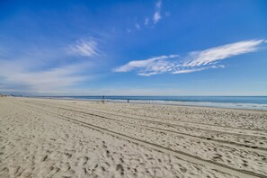 A wide sandy beach with footprints leading to the water, a clear sky, and distant buildings.