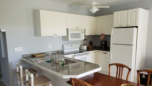 Una cocina con armarios blancos, encimera de granito, microondas, refrigerador y un comedor con sillas de madera.