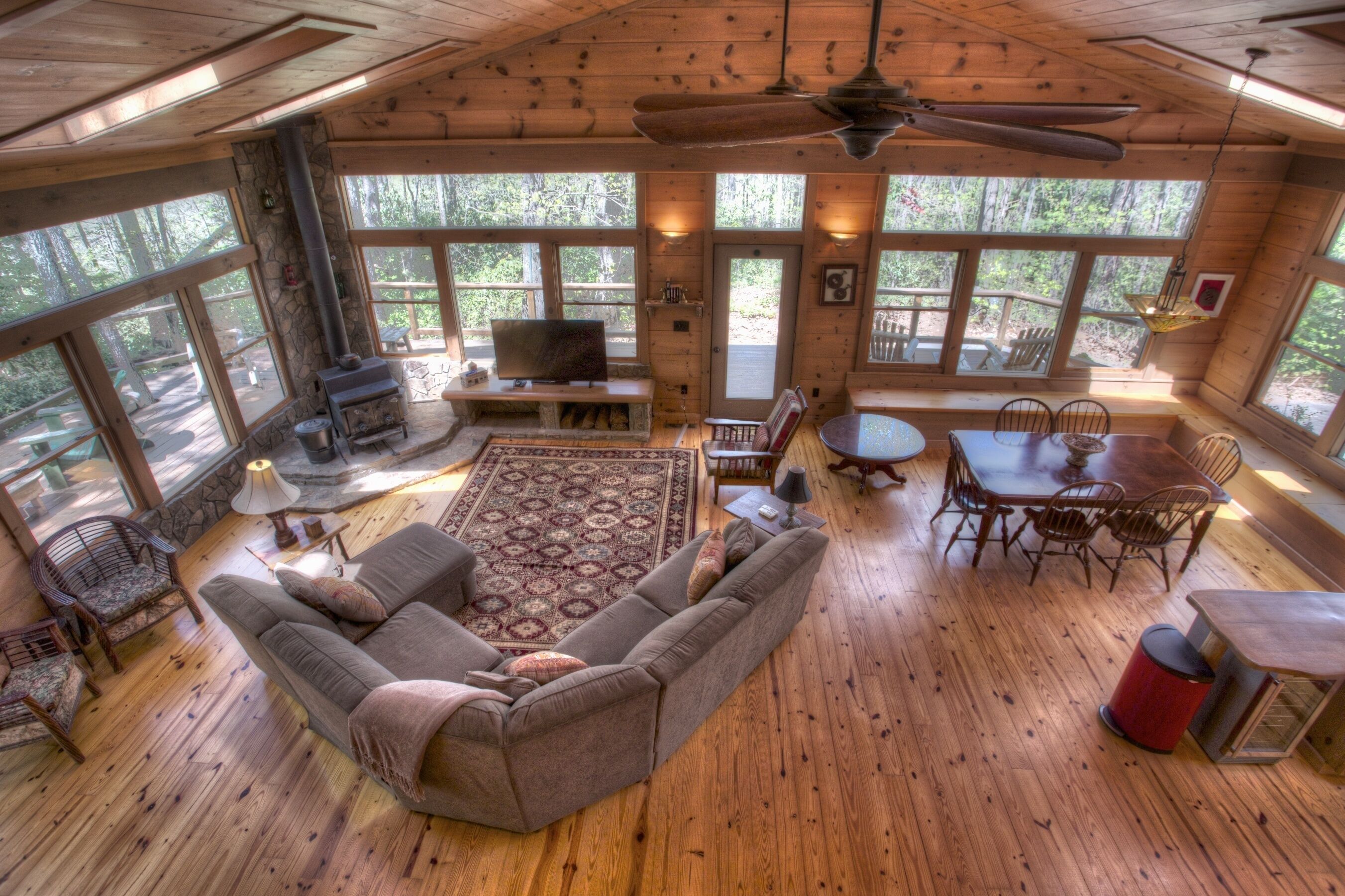 A cozy cabin interior with a stone fireplace, wooden furniture, and large windows offering a view of the forest.
